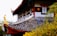 A detailed architectural structure features ornate roof tiles with vibrant hues and intricate patterns. Wooden beams painted in red accentuate the roof, while decorative yellow elements enhance the design. Surrounding foliage, with shades of green and yellow, provides a natural backdrop.