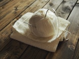 A ball of cream-colored yarn is placed on top of a knitted fabric piece. Circular knitting needles are inserted into the yarn. The setup rests on a wooden surface with a warm, rustic appearance.