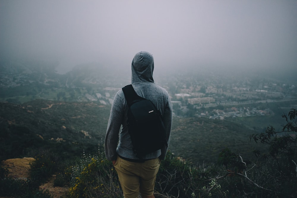 Hombre con sudadera con capucha gris con mochila negra mirando la ciudad desde la montaña