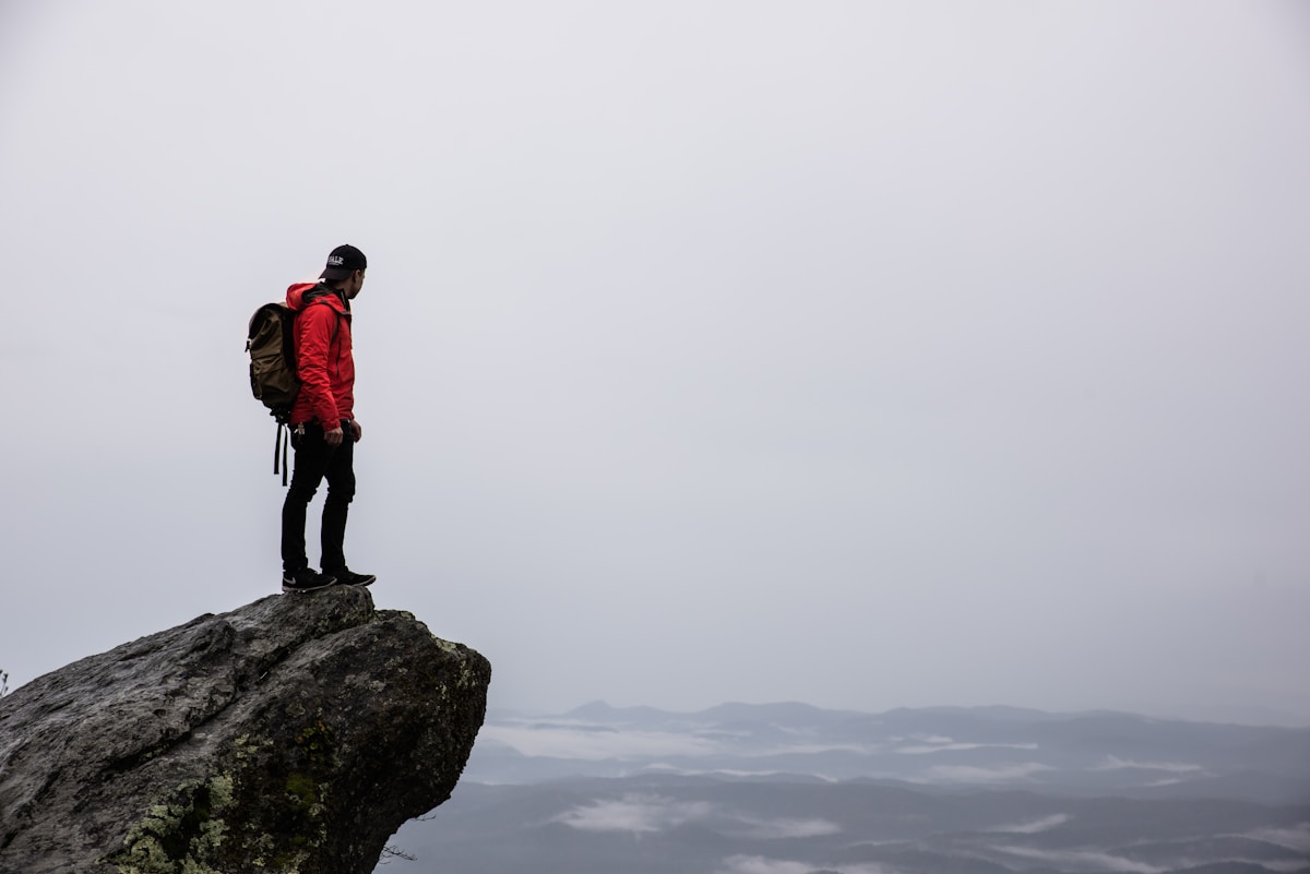 Person wearing a reversed black baseball cap, red parka, black jeans and sneakers and a backpack standing on a cliff looking out at fog covered mountains beyond.