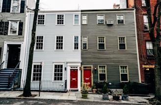 A three-story residential building features a striking split design with the left side painted light blue and the right side dark gray. Each side has a bright red door with a small rectangular window above. Windows are symmetrically aligned across the facade, and a small black metal fence separates the property from the street. Sparse vegetation, including potted plants, is present in front of the building.