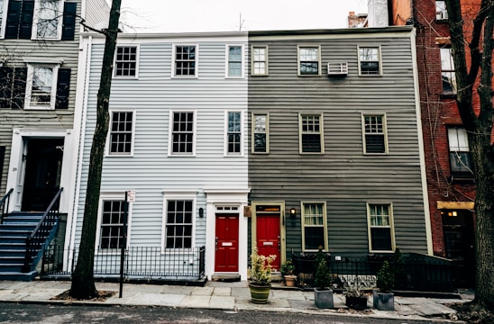 A three-story residential building features a striking split design with the left side painted light blue and the right side dark gray. Each side has a bright red door with a small rectangular window above. Windows are symmetrically aligned across the facade, and a small black metal fence separates the property from the street. Sparse vegetation, including potted plants, is present in front of the building.