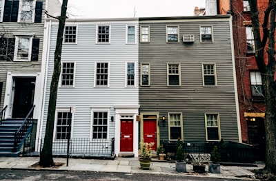 A three-story residential building features a striking split design with the left side painted light blue and the right side dark gray. Each side has a bright red door with a small rectangular window above. Windows are symmetrically aligned across the facade, and a small black metal fence separates the property from the street. Sparse vegetation, including potted plants, is present in front of the building.