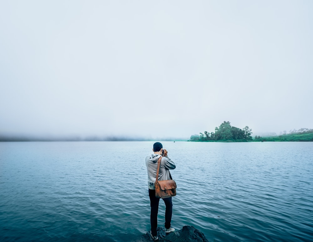 man holding brown leather saddlebag standing on edge of rock over body of water