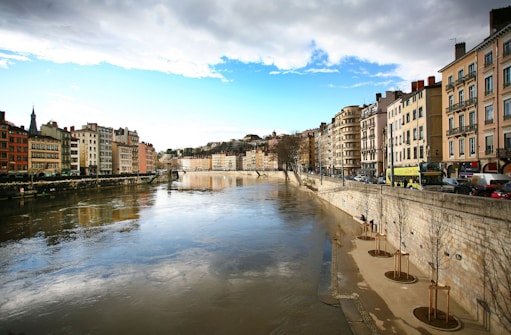 A scenic urban riverfront with multi-story buildings lining both sides of a wide river. The buildings display various architectural styles and colors, ranging from modern to classic. The sky is partly cloudy with some blue patches visible. There are trees along the riverbank, some recently planted and supported with wooden stakes. Cars and a bus travel along the street adjacent to the river.
