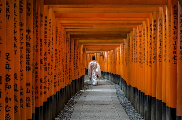 A person dressed in traditional white Japanese clothing walks along a path lined by numerous bright orange torii gates. The gates are densely placed, creating a tunnel-like effect, and each gate is inscribed with black Japanese characters. The pathway is paved with stones and is flanked by small pebbles.
