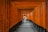 A person dressed in traditional white Japanese clothing walks along a path lined by numerous bright orange torii gates. The gates are densely placed, creating a tunnel-like effect, and each gate is inscribed with black Japanese characters. The pathway is paved with stones and is flanked by small pebbles.