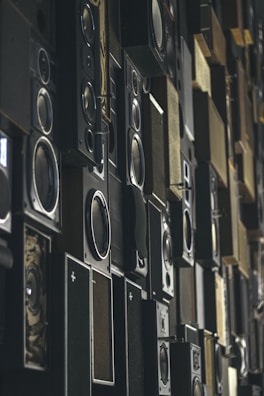 Multiple speaker boxes are arranged in a grid-like pattern, creating a textured and layered visual effect. The speakers vary in size and design, with some displaying intricate details and others having a minimalist appearance.