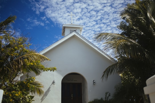 A small white chapel with a cross on top is surrounded by palm trees under a blue sky with scattered clouds. The building has a simple entrance with two outdoor lamps on either side of the door.