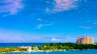 A picturesque view of a coastal area with a clear blue sky and ocean in the background. The foreground features lush greenery and some buildings, including a large, ornate hotel on the right.
