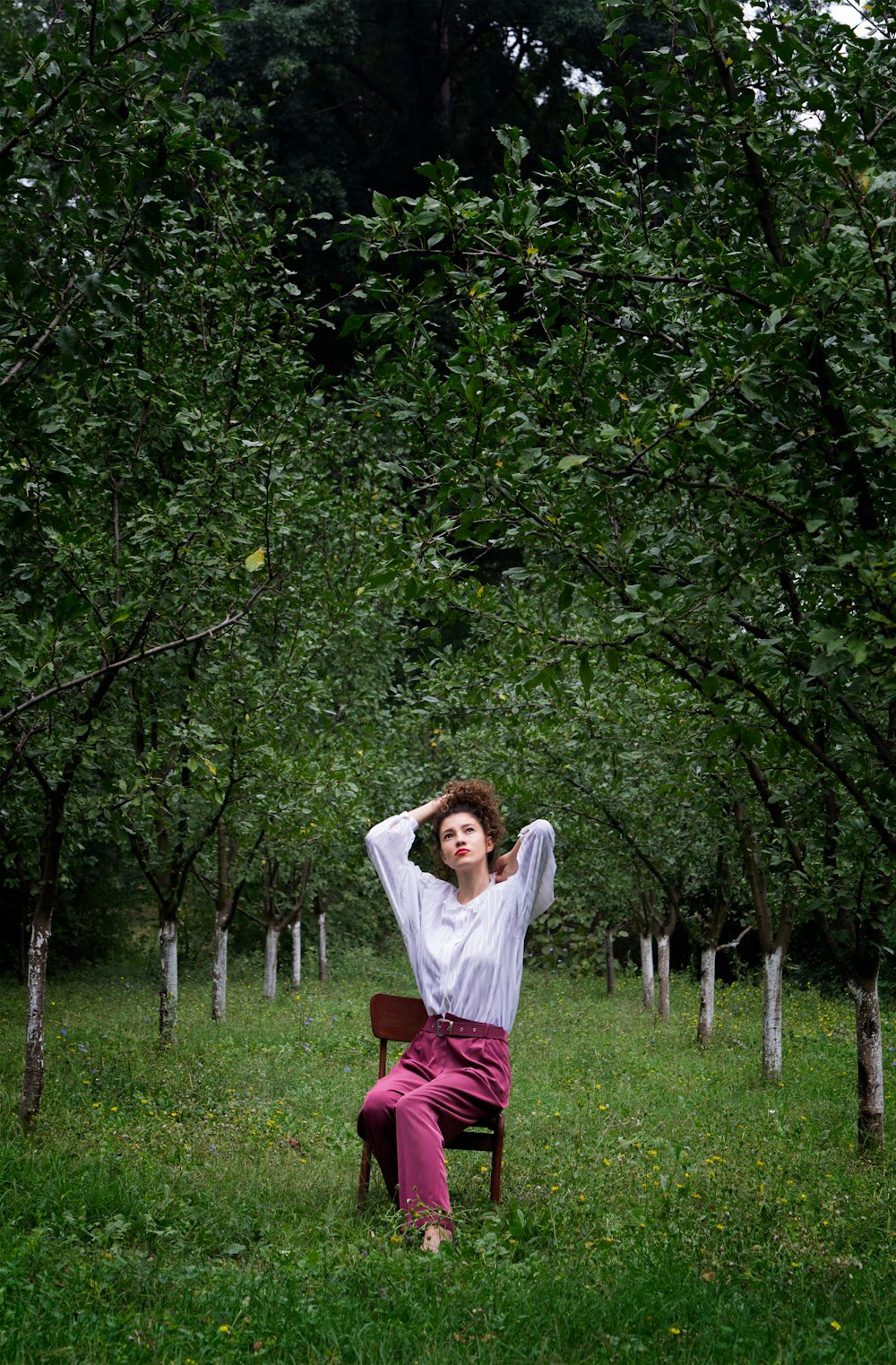 woman sitting on brown armless chair while tying her hair under green trees at daytime