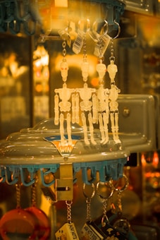 Several skeleton-shaped keychains hang from hooks in a display stand, set against a warm, ambient background. The keychains appear translucent with red eye details, and the display has a blue and clear structure with the word 'WIN' visible.