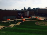 A series of brightly colored orange and black umbrellas are arranged in a pattern on a grassy area. The setting includes a modern brick building with large banners promoting a dance event. There are trees in the background, and the scene appears to be part of a university or campus environment.