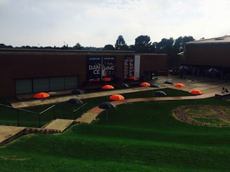 A series of brightly colored orange and black umbrellas are arranged in a pattern on a grassy area. The setting includes a modern brick building with large banners promoting a dance event. There are trees in the background, and the scene appears to be part of a university or campus environment.