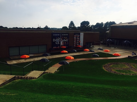 A series of brightly colored orange and black umbrellas are arranged in a pattern on a grassy area. The setting includes a modern brick building with large banners promoting a dance event. There are trees in the background, and the scene appears to be part of a university or campus environment.
