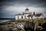 A historic lighthouse stands on a rocky shoreline under overcast skies. Driftwood is scattered across the rocky beach, and muted grass lines the edge leading up to the building. The lighthouse is white with a red roof and features a prominent lantern room.