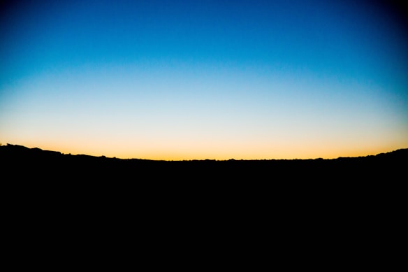 A vibrant horizon where the deep blue sky transitions gracefully into a warm yellow and orange hue, with a dark, silhouetted landscape stretching across the lower part of the image.