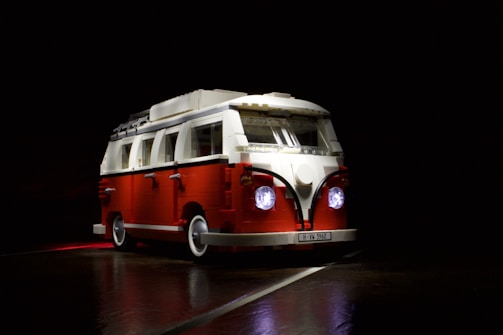 A miniature van model made from toy bricks, primarily red and white, with two illuminated headlights. The vehicle is portrayed against a dark background, highlighting its detailed features including side mirrors, a luggage rack, and a license plate reading 'H-VW 1962'.