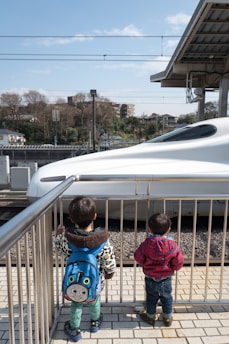 Two young children stand on a platform looking at a high-speed train passing by. One child wears a blue backpack featuring a cartoon character, while the other is dressed in a red checkered jacket. The scene is bright and sunny with a clear blue sky, and trees and buildings can be seen in the background.