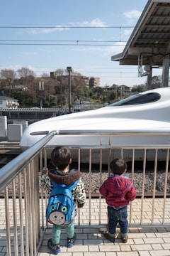 Two young children stand on a platform looking at a high-speed train passing by. One child wears a blue backpack featuring a cartoon character, while the other is dressed in a red checkered jacket. The scene is bright and sunny with a clear blue sky, and trees and buildings can be seen in the background.