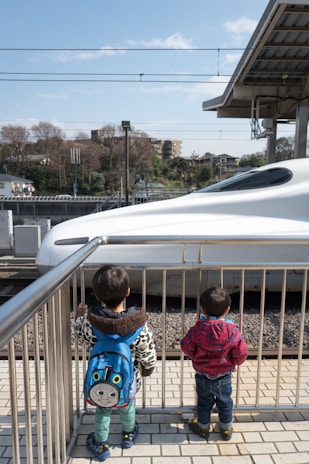 Two young children stand on a platform looking at a high-speed train passing by. One child wears a blue backpack featuring a cartoon character, while the other is dressed in a red checkered jacket. The scene is bright and sunny with a clear blue sky, and trees and buildings can be seen in the background.