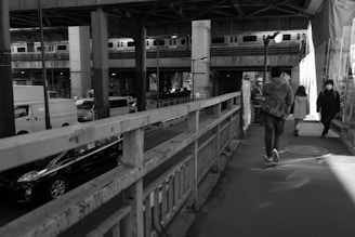 Urban street scene beneath an overpass, featuring pedestrians walking on the sidewalk and vehicles like buses and taxis on the road. Above, a train moves along elevated tracks. Construction workers and pedestrians are present, some wearing masks.