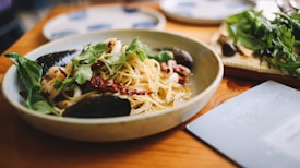 A bowl of pasta garnished with fresh herbs and seafood, including mussels and squid, is placed on a wooden table. Next to it, there is a plate of greens and a menu or booklet.