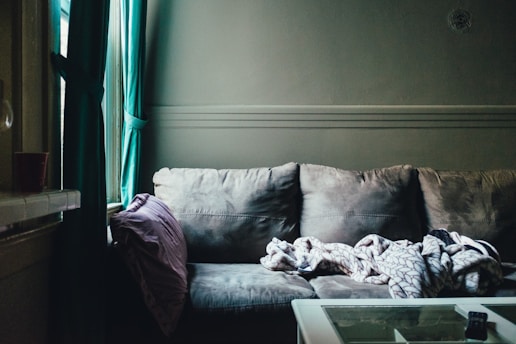 A cozy, slightly dim living room corner with a plush gray sofa. A folded, patterned blanket rests on the seat, accompanied by a purple pillow. Teal curtains hang on a window beside the couch, casting a soft natural light into the room. A small part of a white table with a remote control is visible in the foreground.