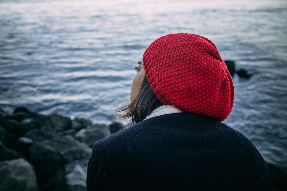 Frau in schwarzer Jacke und roter Strickmütze steht tagsüber am felsigen Ufer