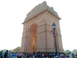 A large stone archway monument stands prominently in an open space. The structure is detailed with carvings and inscriptions, with several flags fluttering nearby. People are gathered around the monument, indicating a busy and possibly a tourist area. A street lamp is positioned to the right, complementing the historical ambiance.