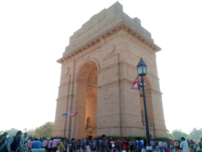 A large stone archway monument stands prominently in an open space. The structure is detailed with carvings and inscriptions, with several flags fluttering nearby. People are gathered around the monument, indicating a busy and possibly a tourist area. A street lamp is positioned to the right, complementing the historical ambiance.