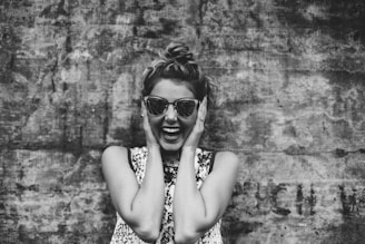 A joyful woman with sunglasses smiles broadly while holding her face with both hands. Her hair is tied up in a stylish bun, and she is wearing a patterned sleeveless top. The background is a textured concrete wall, giving the image a rustic, urban feel.
