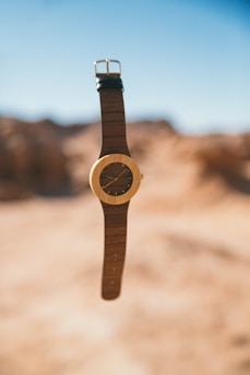 A wooden wristwatch with a leather band is centered against a blurred desert landscape. The watch face is round with a minimalist design, featuring no numbers and simple gold hands. The desert background is hazy, showcasing shades of brown and blue sky, providing a serene and calm setting.