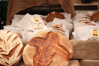 Various artisan breads placed on a burlap-covered table, with two distinct loaves prominently displayed in the foreground. Each loaf has a unique pattern or scoring on the crust and is surrounded by paper bags labeled with different types of bread, such as 'Red Fife Glenn Boule' and 'Sonora Country Blonde'.