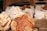 Various artisan breads placed on a burlap-covered table, with two distinct loaves prominently displayed in the foreground. Each loaf has a unique pattern or scoring on the crust and is surrounded by paper bags labeled with different types of bread, such as 'Red Fife Glenn Boule' and 'Sonora Country Blonde'.