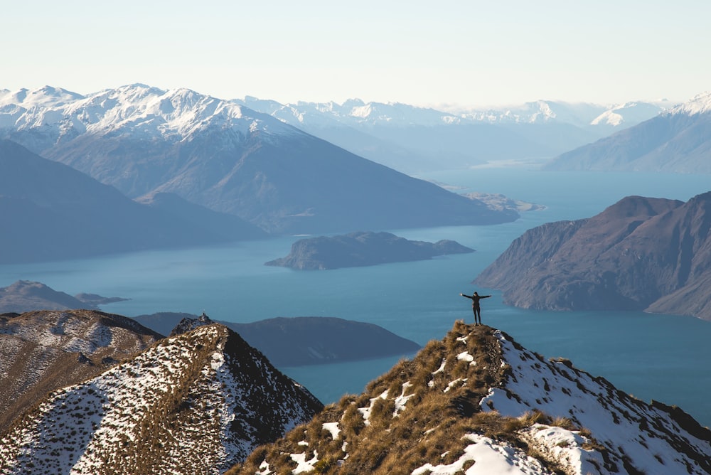 Person stands on grassy snow-covered summit overlooking water with arms outstretched