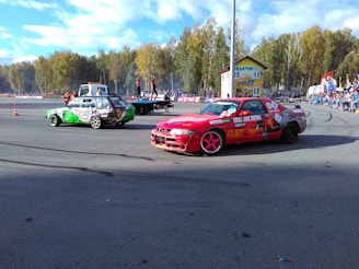 Two customized cars are on a racetrack, one red with a striking design and the other a green and white vehicle. People are observing from the sidelines, and a platform with individuals is visible in the background. Trees and a building are present, suggesting an outdoor environment.