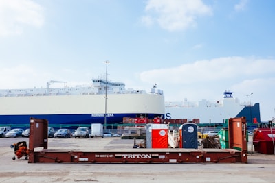 A large cargo ship is docked at a port. In the foreground, there is a brown metal platform with the label 'TRITON'. Behind the platform, there are two portable toilets, one red and one blue, along with other miscellaneous items. Several cars are parked on the port, and metal containers are stacked in the background.