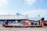 A large cargo ship is docked at a port. In the foreground, there is a brown metal platform with the label 'TRITON'. Behind the platform, there are two portable toilets, one red and one blue, along with other miscellaneous items. Several cars are parked on the port, and metal containers are stacked in the background.