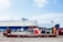 A large cargo ship is docked at a port. In the foreground, there is a brown metal platform with the label 'TRITON'. Behind the platform, there are two portable toilets, one red and one blue, along with other miscellaneous items. Several cars are parked on the port, and metal containers are stacked in the background.