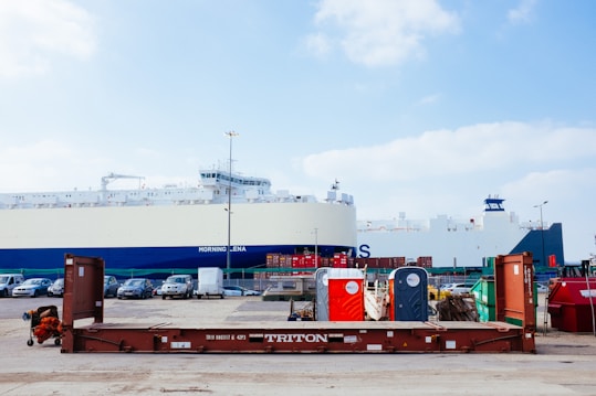 A large cargo ship is docked at a port. In the foreground, there is a brown metal platform with the label 'TRITON'. Behind the platform, there are two portable toilets, one red and one blue, along with other miscellaneous items. Several cars are parked on the port, and metal containers are stacked in the background.