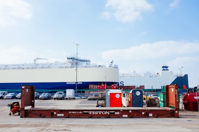 A large cargo ship is docked at a port. In the foreground, there is a brown metal platform with the label 'TRITON'. Behind the platform, there are two portable toilets, one red and one blue, along with other miscellaneous items. Several cars are parked on the port, and metal containers are stacked in the background.