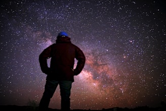 A person stands silhouetted against a vibrant, star-filled night sky, with the Milky Way galaxy clearly visible. The scene captures the vastness of the universe and the smallness of the human figure in comparison, evoking a sense of wonder and awe.
