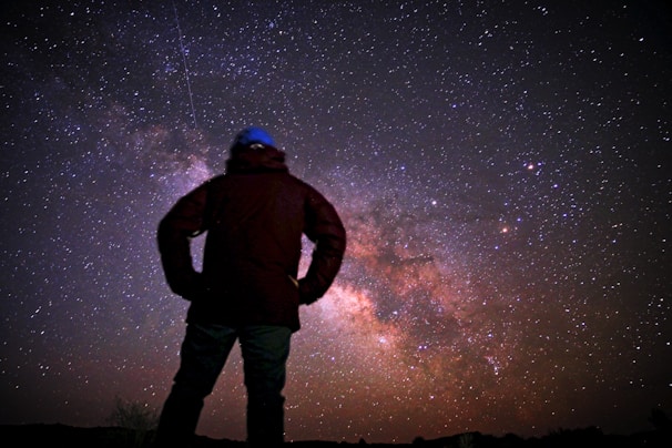 A person stands silhouetted against a vibrant, star-filled night sky, with the Milky Way galaxy clearly visible. The scene captures the vastness of the universe and the smallness of the human figure in comparison, evoking a sense of wonder and awe.