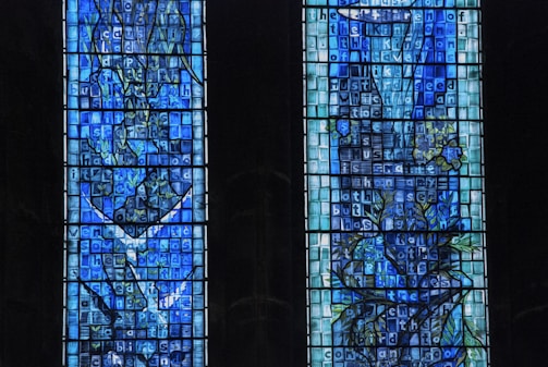 Two tall stained glass windows featuring intricate patterns and designs with various shades of blue and green. The glass panes include text inscribed on them, along with abstract shapes resembling foliage. The overall structure is symmetric, and the artistry demonstrates a mix of geometric and organic elements.
