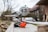 A pair of orange and black protective earmuffs placed on a wooden surface, with a blurred circular saw in the background. The setting appears to be outdoors, perhaps in a backyard or workshop area. The focus is on the earmuffs, with the woodworking tool and natural surroundings softly out of focus.
