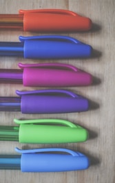 Six pens with colorful caps are arranged parallel to each other on a wooden surface. The caps are colored in orange, blue, pink, purple, green, and light blue respectively.