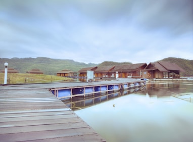 A tranquil lakeside scene with wooden lodges on stilts extending over calm water. The lodges feature sloping roofs and are surrounded by lush green hills under a cloudy sky. A wooden dock leads to the lodges, enhancing the serene atmosphere.