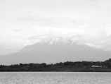 A large mountain dominates the center of the scene, partially covered by clouds and with snow visible on its peak. In the foreground, a large body of water spreads out, and beyond it, a strip of lush greenery with trees lines the shore.