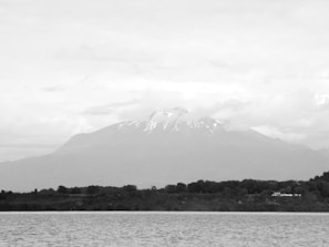 A large mountain dominates the center of the scene, partially covered by clouds and with snow visible on its peak. In the foreground, a large body of water spreads out, and beyond it, a strip of lush greenery with trees lines the shore.